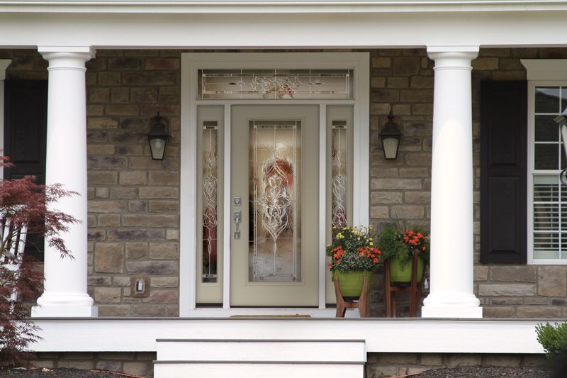 Newly installed front door on light tan brick home with front porch with white columns to either side of front steps. Doors is light tan/gray with decorative glass and decorative sidelites and transom windows surrounding.