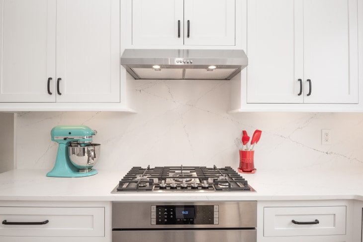 Stainless steel gas stove, oven, and range hood with white shaker cabinets surrounding, white stone countertops and backsplash. Teal KitchenAid mixer to left of stove. Red cooking utensils holder to right of stove.