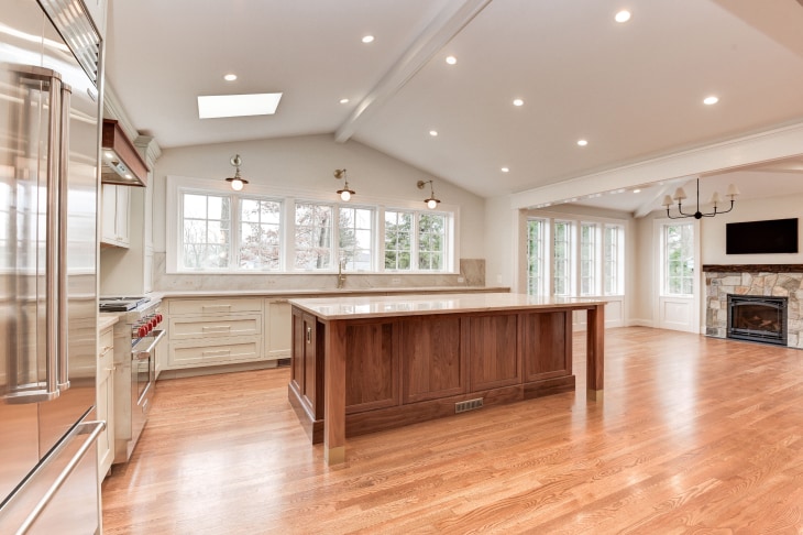 Large open-concept kitchen remodel with wood floors, vaulted white ceiling with recessed lighting, large woodgrain island in middle, white cabinets against far wall. Row of double-hung windows with grids above kitchen countertop. living area in background, to the right, with stone fireplace.