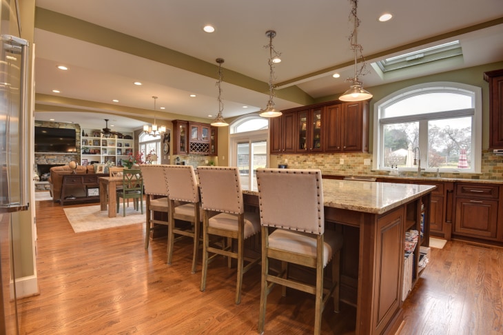 Open-plan kitchen leading to living room area in background. Wood floors, wood cabinets, long island with stool seating. 