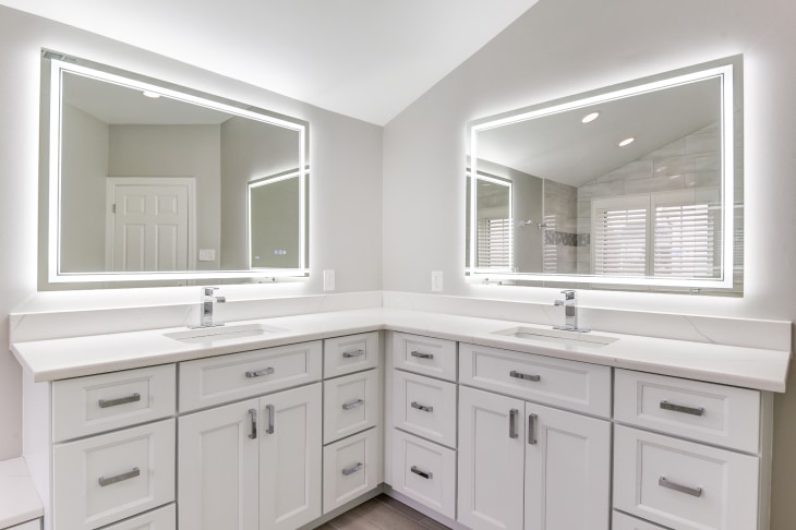 Corner of bathroom remodel, with dual vanity, white cabinets and countertop, light gray walls, and smart mirror above each sink with LED white light bordering each.