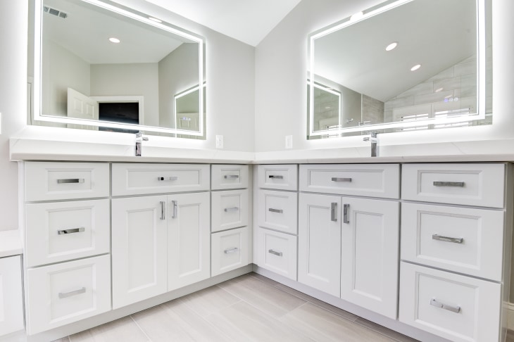 White bathroom remodel with dual vanities, large lighted mirrors over each sink, and white wood-look tile floor.